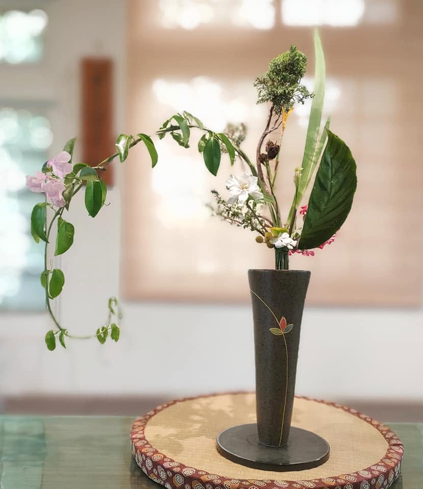 明亮室內的立華新風體作品。細長黑色花器中，枝葉向左延伸，搭配淡紫色與白色花朵、大片綠葉，置於圓形台座上。